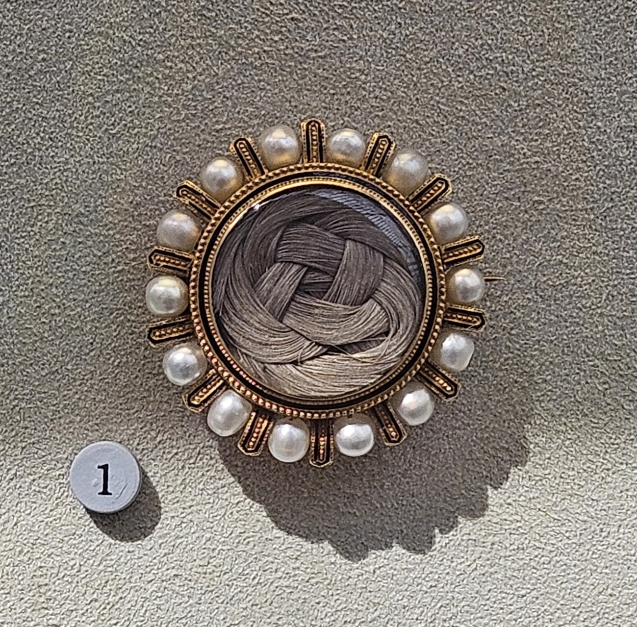 A circular brooch surrounded by gold and inlaid pearls, with an intricately braided lock of hair in the center.