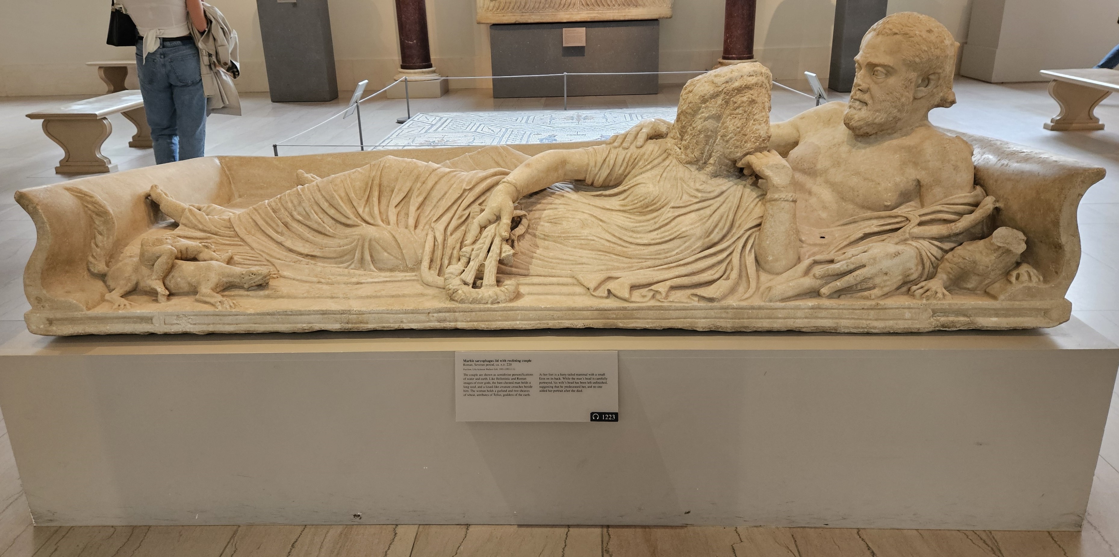 A marble sculpture of a man and a woman reclining while embracing each other. The man's face is detailed, whereas the woman's remains a blank slab of marble.